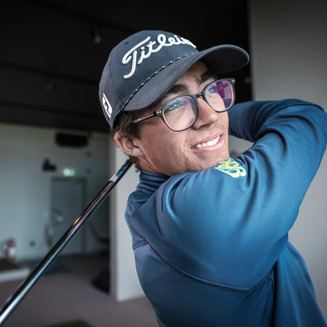 A golfer wearing a Titleist cap and glasses smiles while holding a club over his shoulder, posing inside a golf facility.