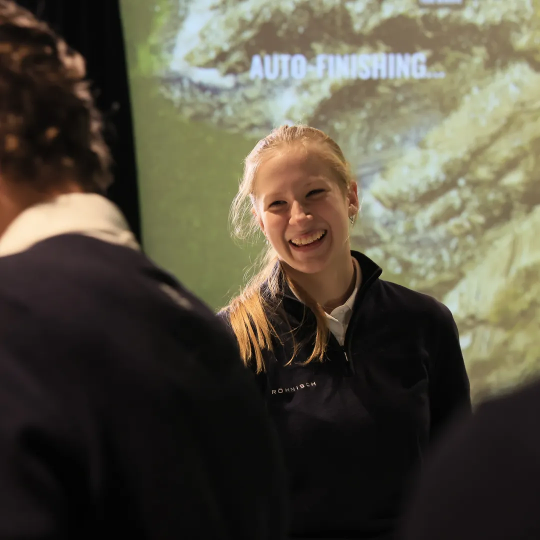 Smiling person in a navy jacket stands in front of an abstract green background, interacting with others in a dimly lit environment.