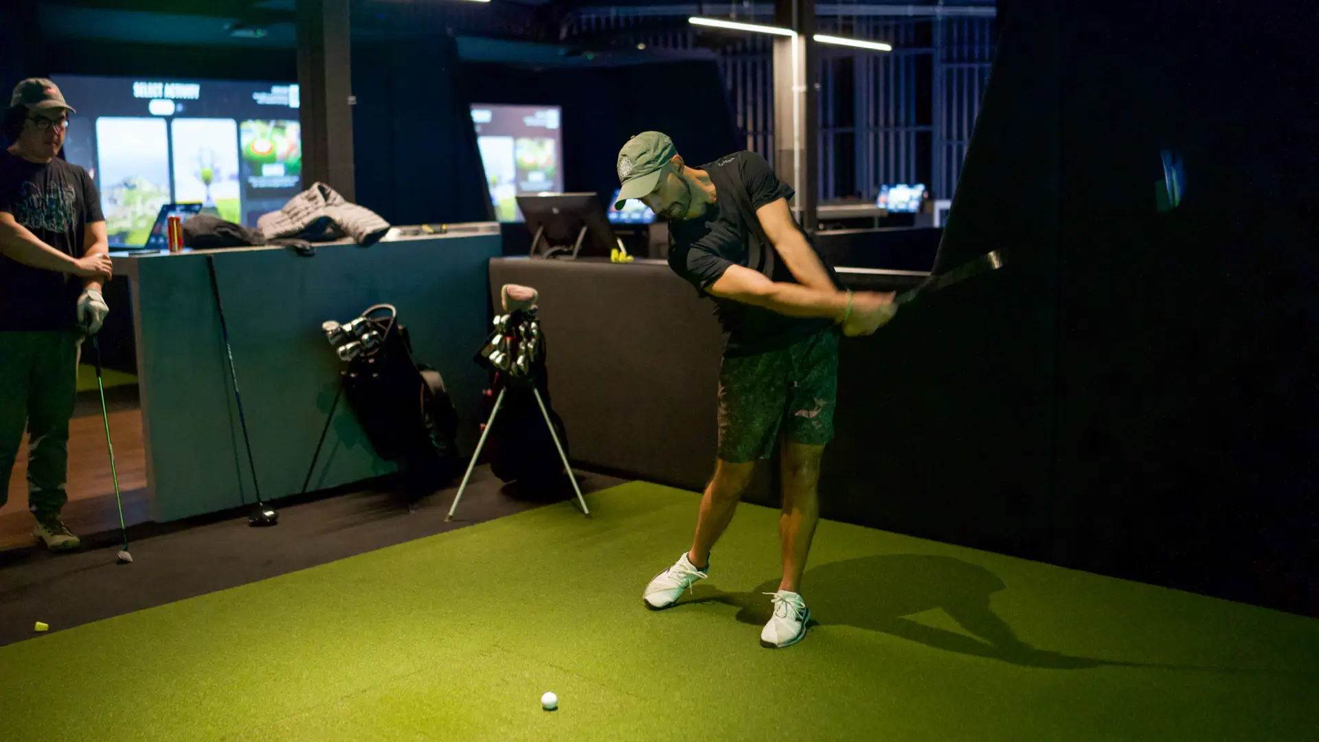 Person swinging a golf club indoors on a green mat, watched by another, with golf equipment in the background.