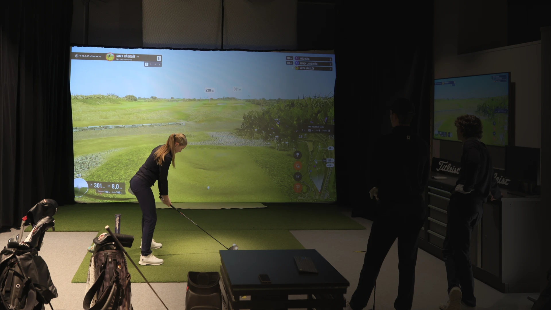 A woman is preparing to swing a golf club in an indoor golf simulator, with a projection of a golf course on the screen.