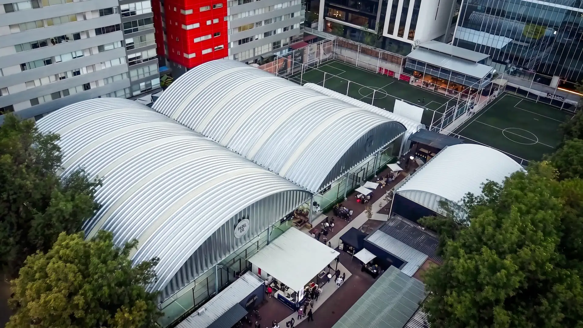Aerial view of curved-roof buildings and a soccer field, surrounded by trees. People gather under canopies beside the structures.