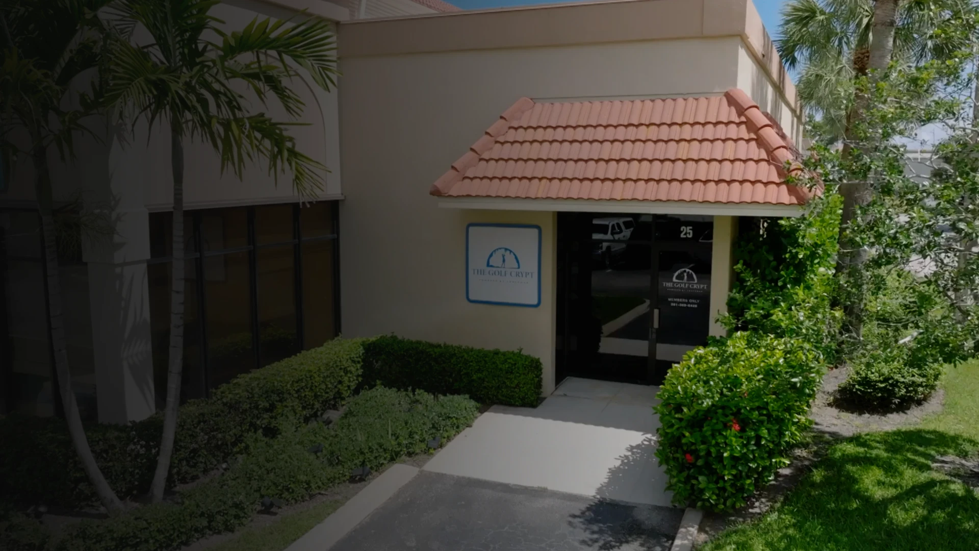 Beige office entrance with red tile awning, glass door and "The Golf Crypt" sign, flanked by palm trees and trimmed green shrubs.