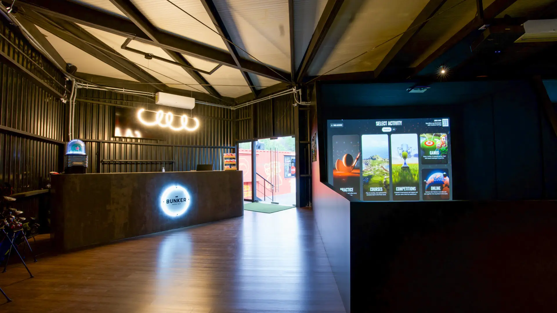 Modern indoor virtual golf center with dark walls, a glowing sign, and a screen displaying activity options. An open door leads outside.