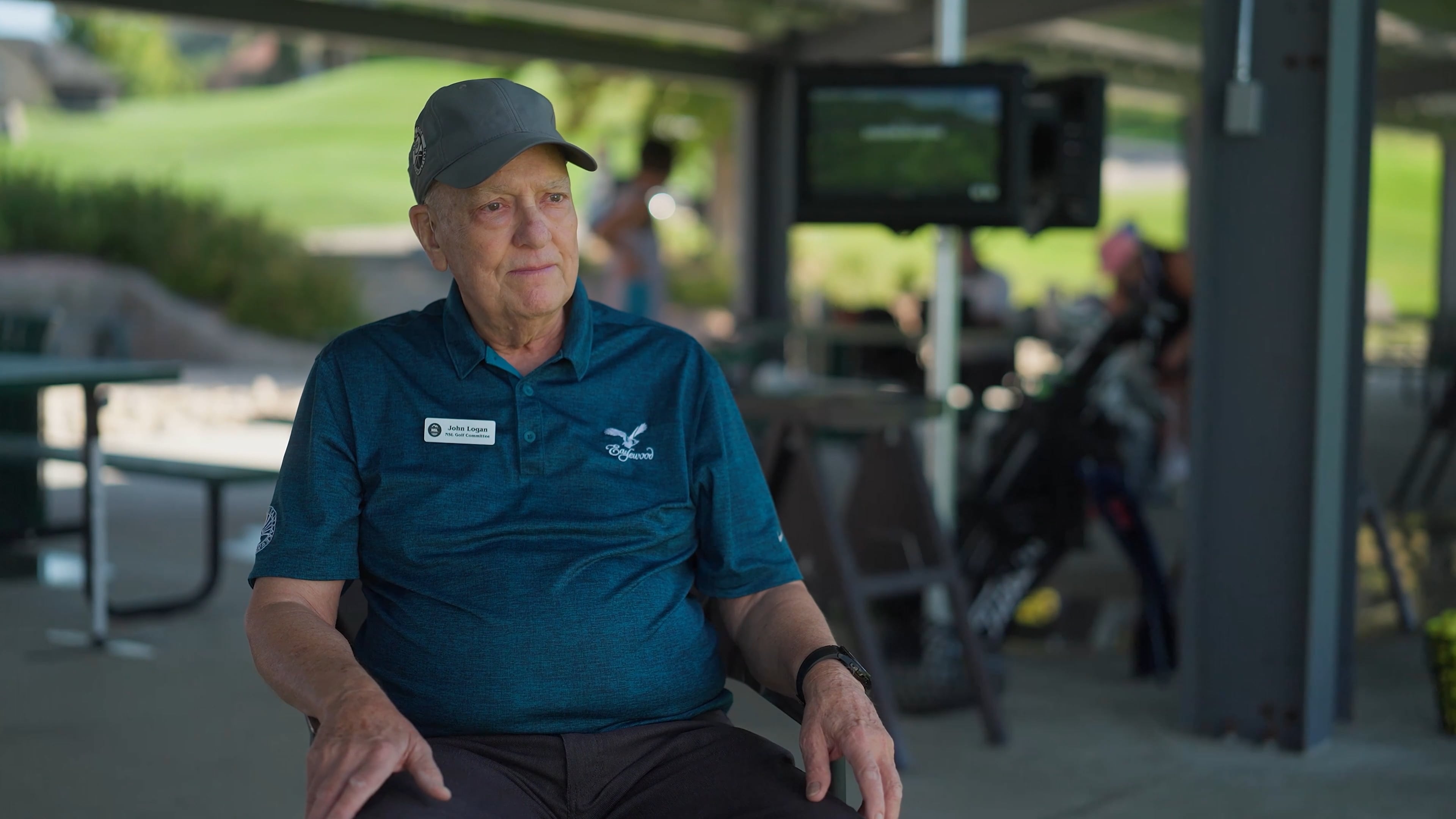 Elderly man in a golf shirt and cap sits outdoors in a shaded area, with a golf course visible in the background.