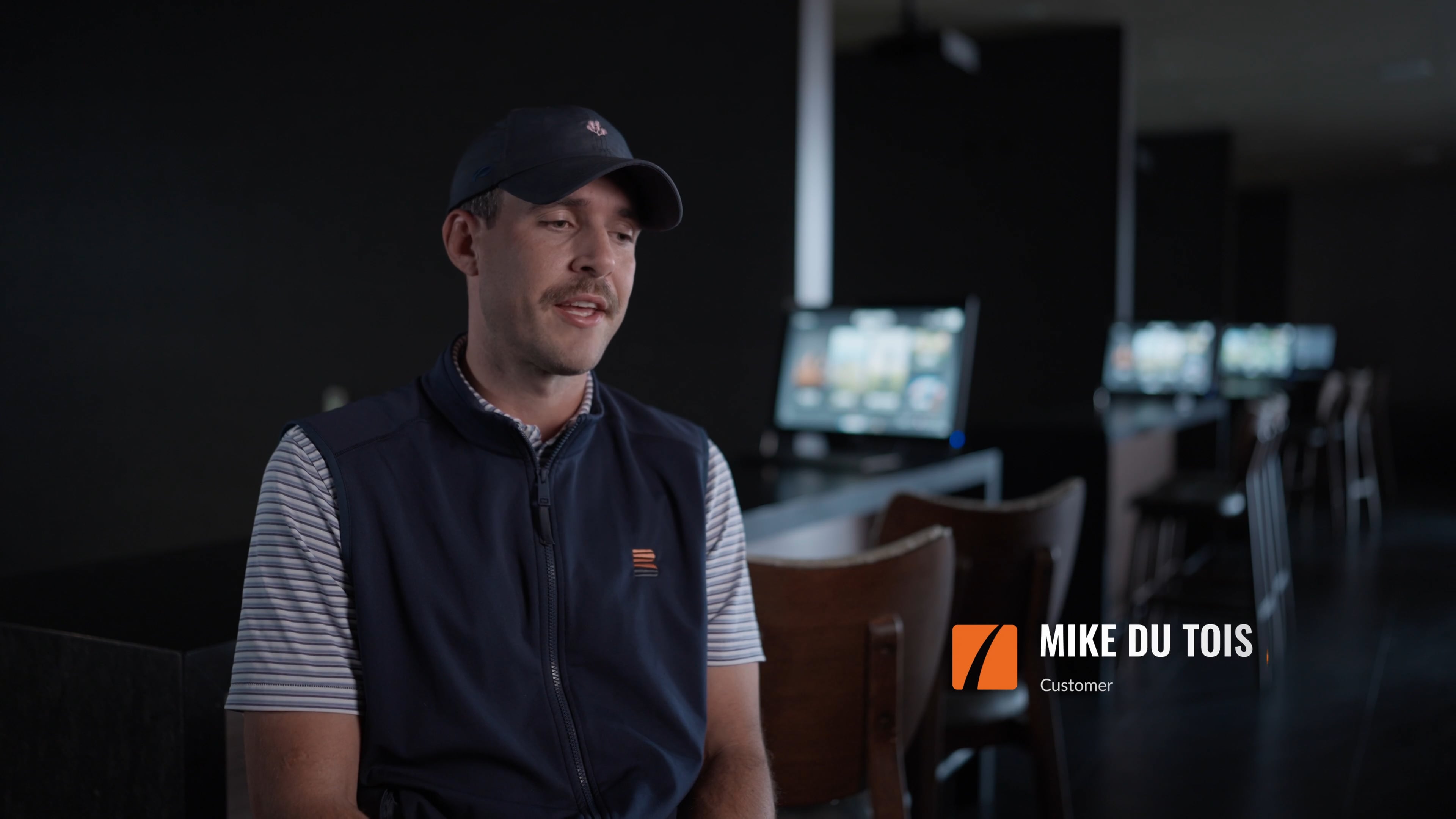 A man wearing a cap and vest is seated, speaking in a dimly lit room. Text on screen says, "Mike Du Tois, Customer."