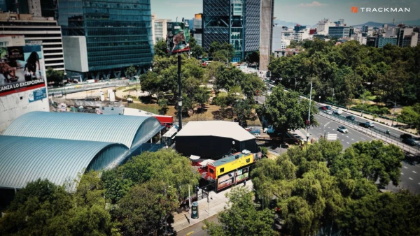 Aerial view of an urban area with modern buildings, greenery, and roads. A curved-roof structure and a billboard stand out among the trees.