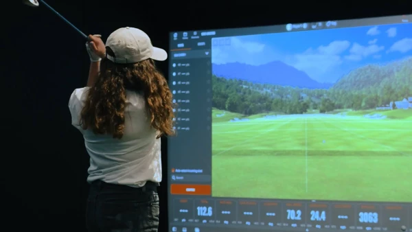 A person with long hair and a cap swings a golf club indoors, facing a virtual golf course projected on a screen.