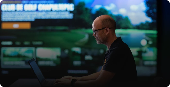 Man using a laptop with a golf game displayed on a large screen in the background.