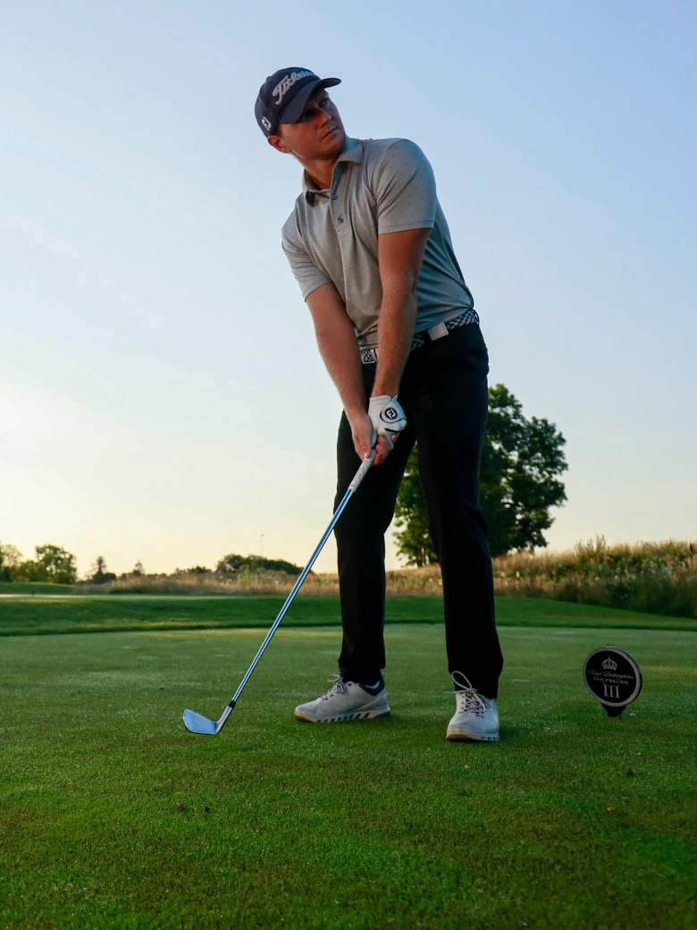 Golfer in a cap and gray shirt preparing to swing on a sunset golf course, with trees and a horizon in the background.