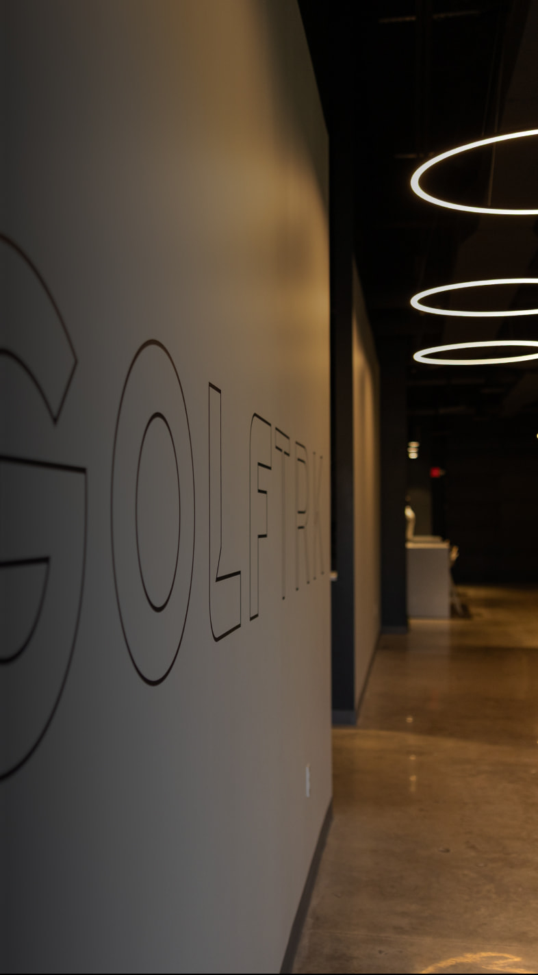 Dimly lit hallway with "GOLFTRK" on the wall, circular ceiling lights, and a distant white reception desk.