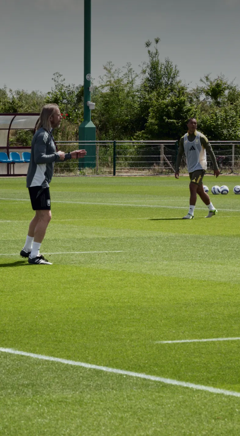 Two soccer players practice on a grassy field, with one preparing to kick a ball. A bench and trees are in the background under a clear sky.