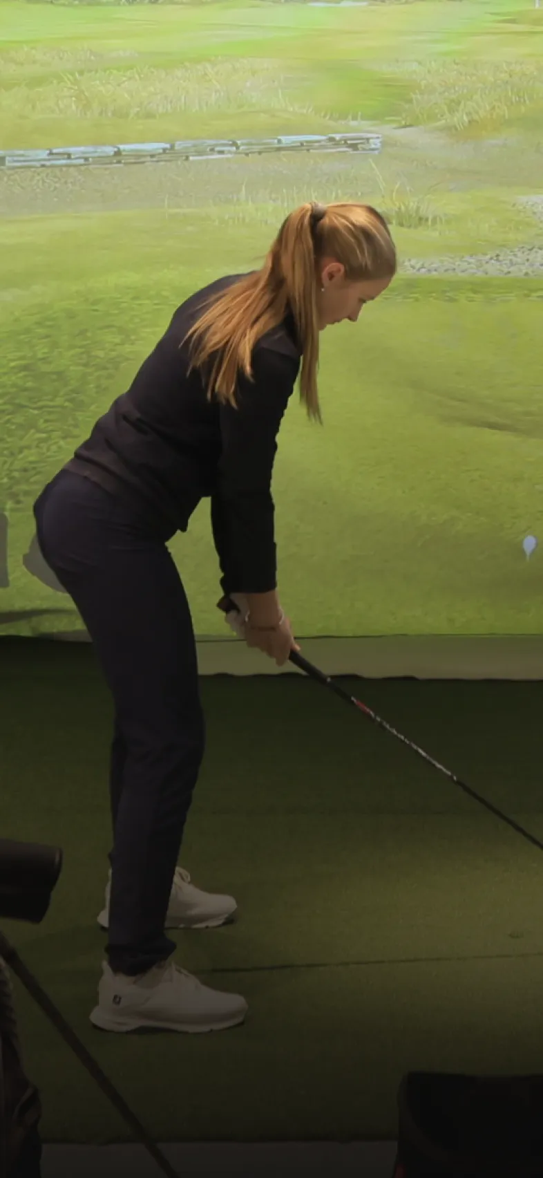 A person with long hair is poised to swing a golf club indoors, facing a virtual golf course screen.