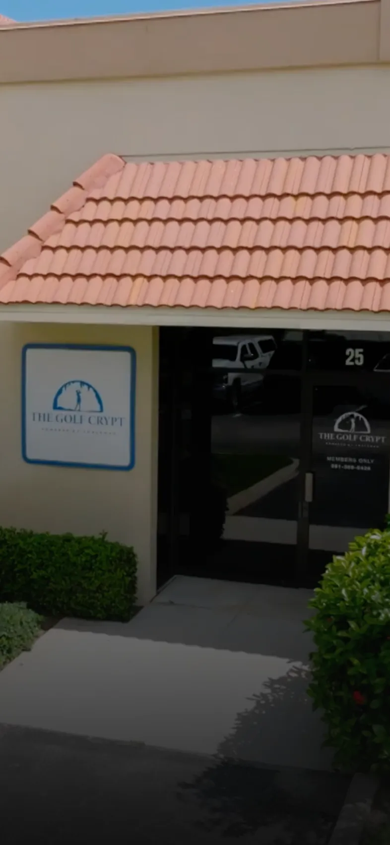 Storefront entrance for The Golf Crypt under a terracotta-tiled awning, dark glass doors and trimmed green shrubs.
