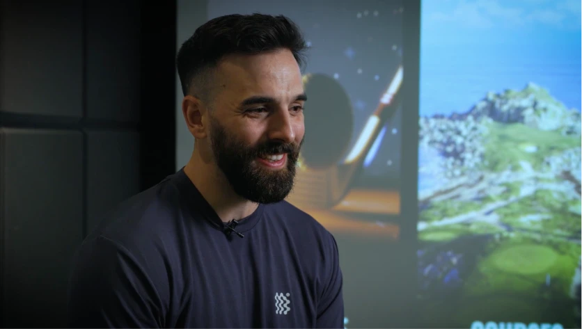 Man with a beard wearing a dark shirt, smiling in a dimly lit room with a golf-themed background.