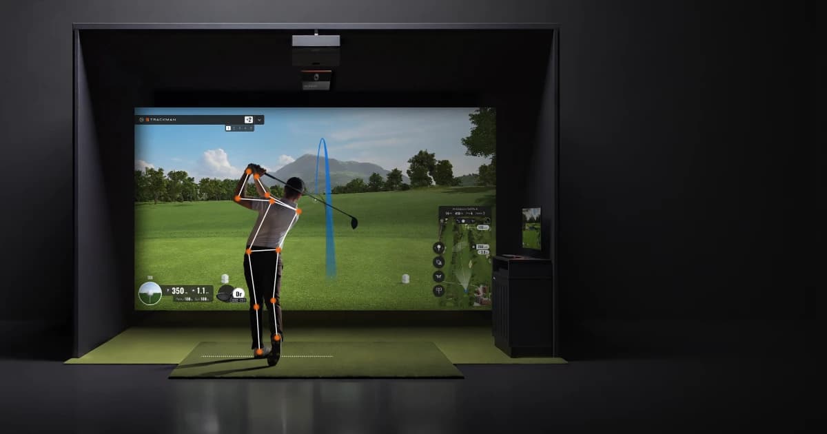 A person swings a golf club in an indoor virtual golf simulator, displaying trajectory and data on a large screen.