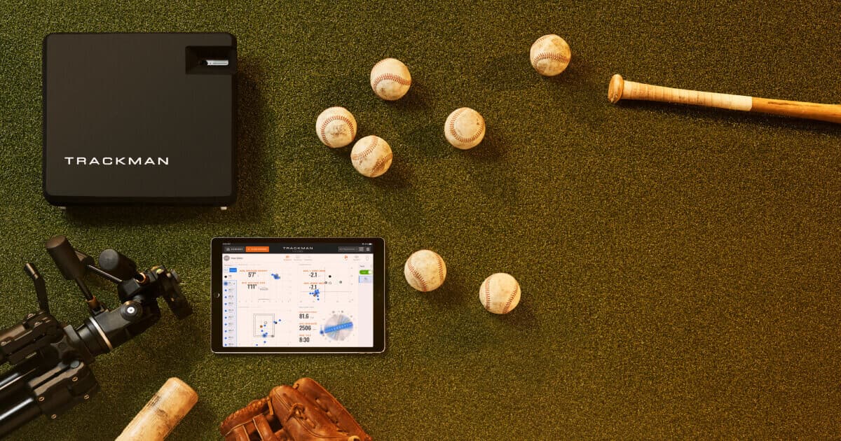 Trackman Baseball equipment on grass: Trackman device, tablet displaying data, camera, wooden bat, glove, and several baseballs.