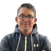 Man with glasses, wearing a dark Under Armour jacket, stands against a white background, arms crossed, smiling slightly.