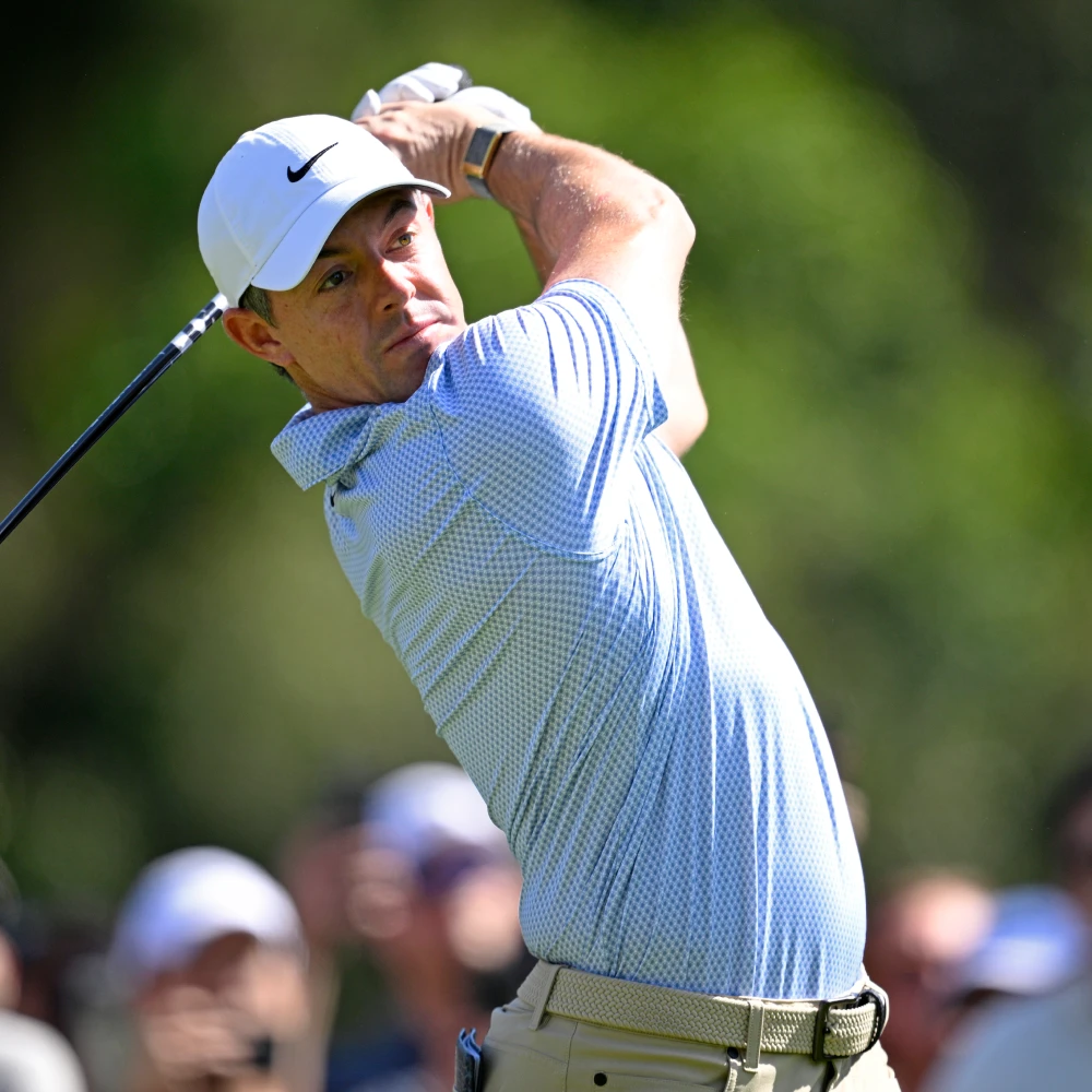 Golfer swinging a club in a blue shirt and white cap, focused, with a blurred background of trees and spectators.