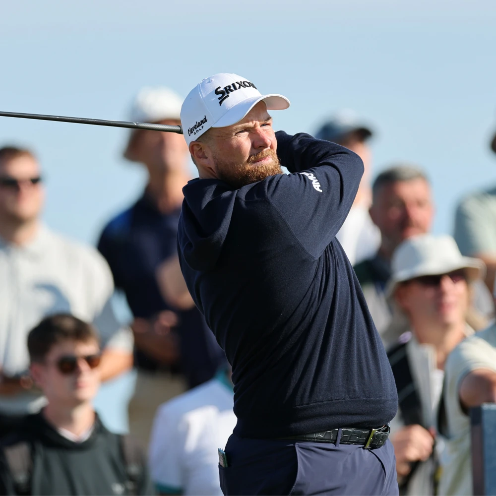 Golfer mid-swing on a sunny day, wearing a white cap and navy shirt. A crowd watches in the background.