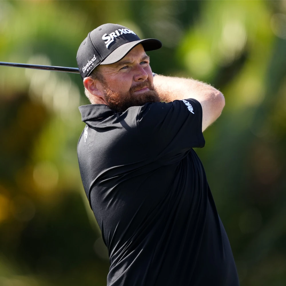 Golfer swinging a club, wearing a black polo and cap, with a focused expression, surrounded by a blurred green background.