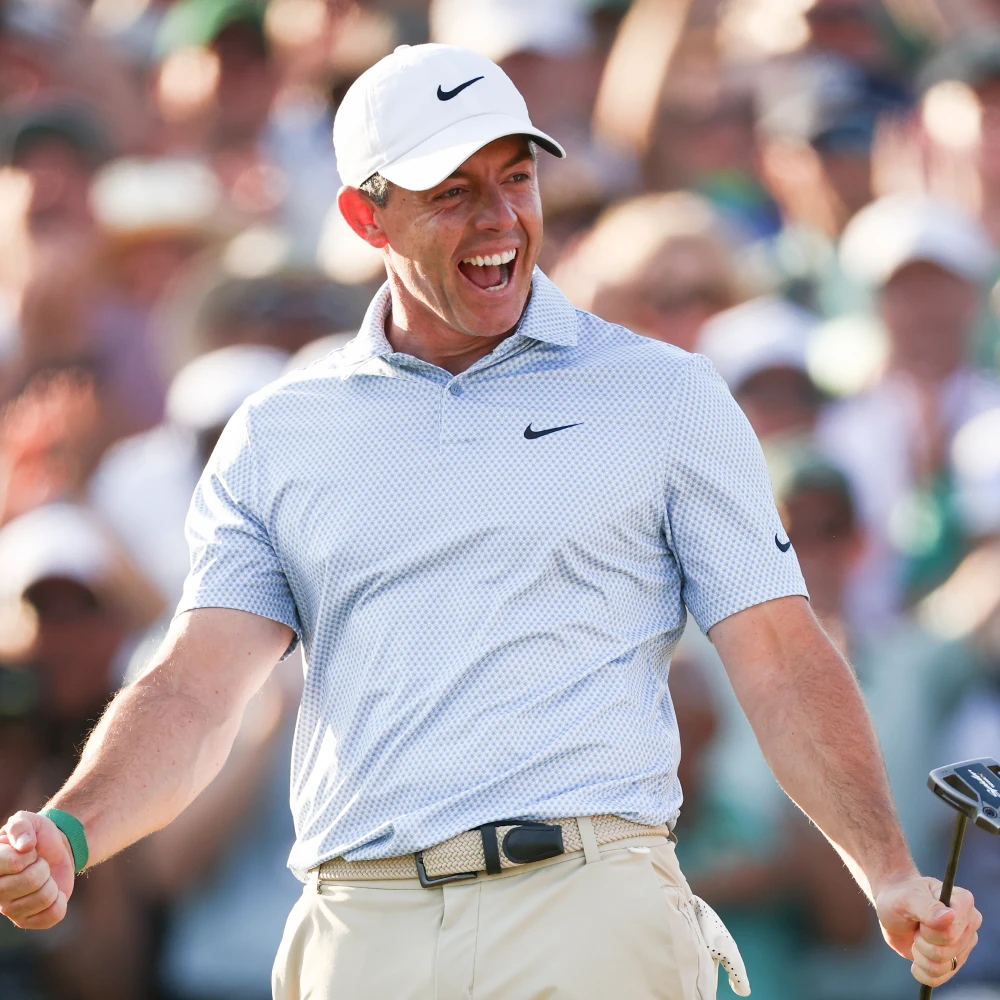 Golfer in a white cap and checkered shirt celebrates with a joyful expression, holding a golf club, surrounded by a cheering crowd.