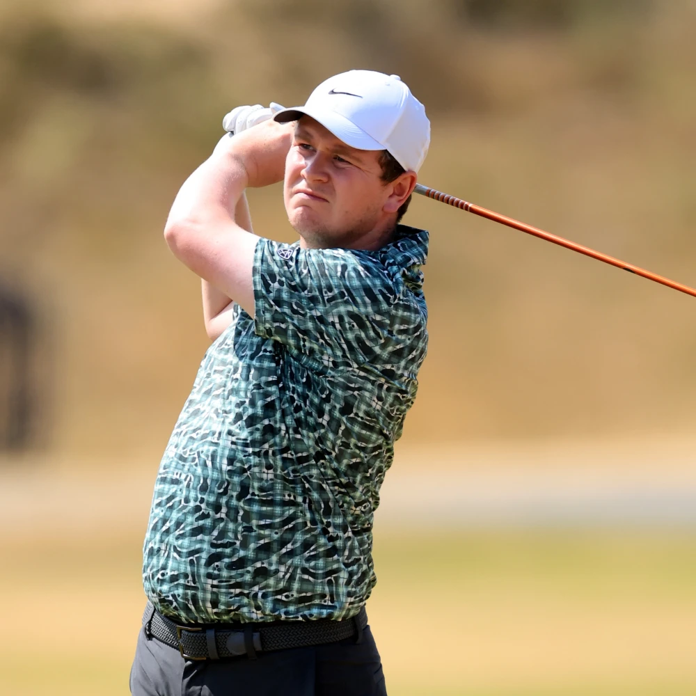 Golfer wearing a white cap and patterned shirt swings a club on a sunny day.