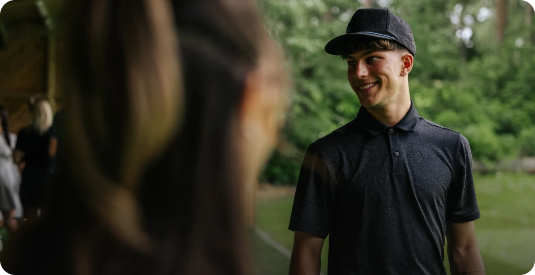 A young man in a dark shirt and cap smiles outdoors, while a woman in the foreground listens, blurred in focus.