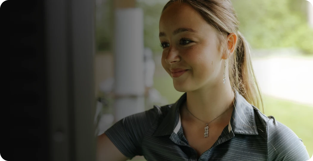 Woman smiling, looking at a screen outdoors, wearing a collared shirt and necklace, with a blurred background of greenery.
