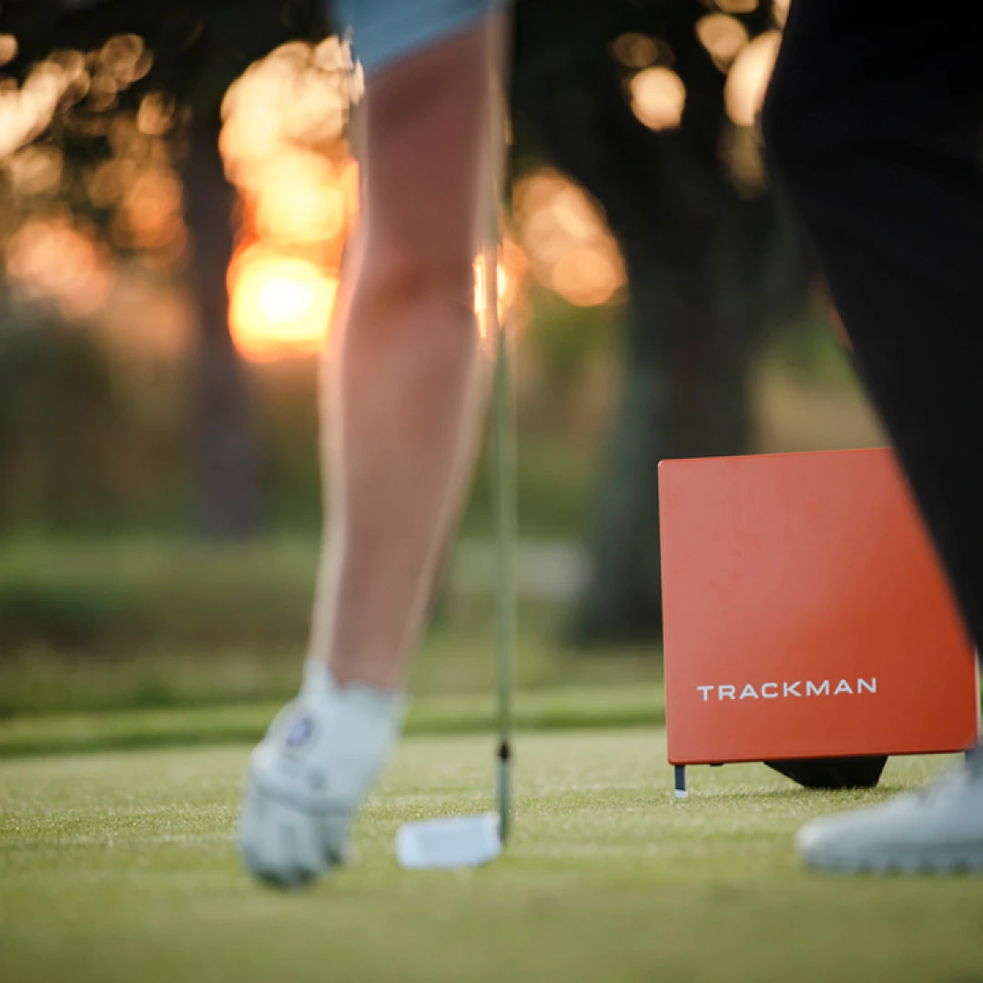 Golfer putting on green at sunset, with an orange Trackman device nearby.
