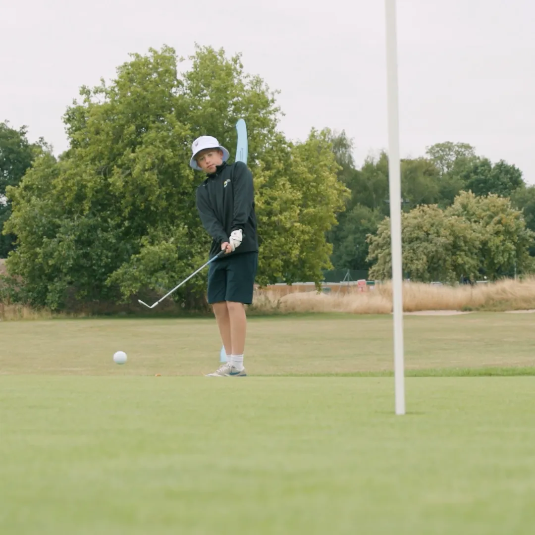 Person in a hat and shorts playing golf, swinging a club near the putting green with trees in the background.