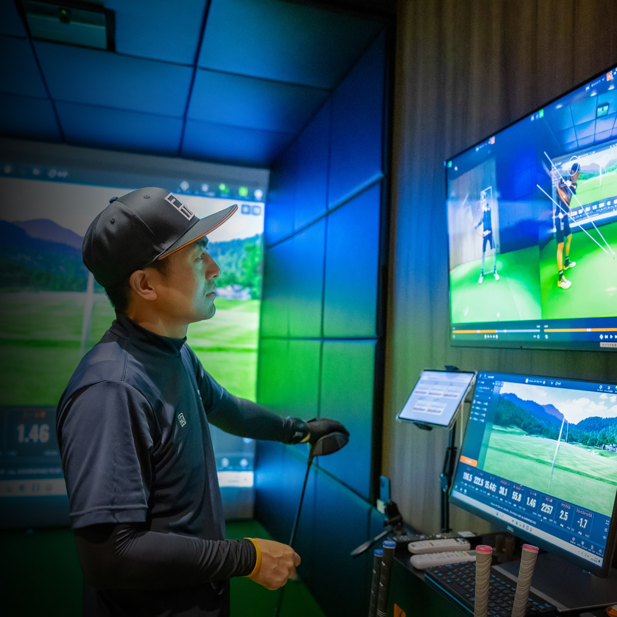 A person in a golf simulator room analyzes their swing on multiple screens, holding a golf club and wearing a cap and dark sportswear.
