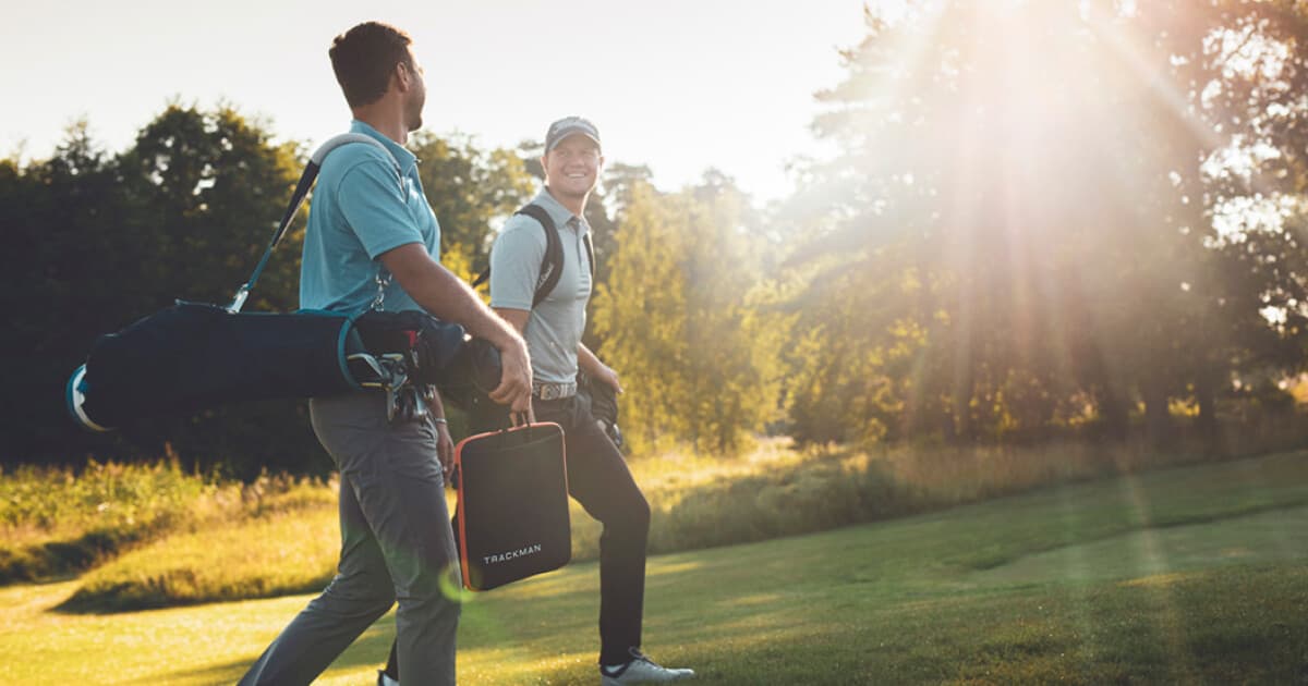 Two people walking on a sunny golf course, carrying golf bags. One person smiles at the other, with trees and sunlight in the background.
