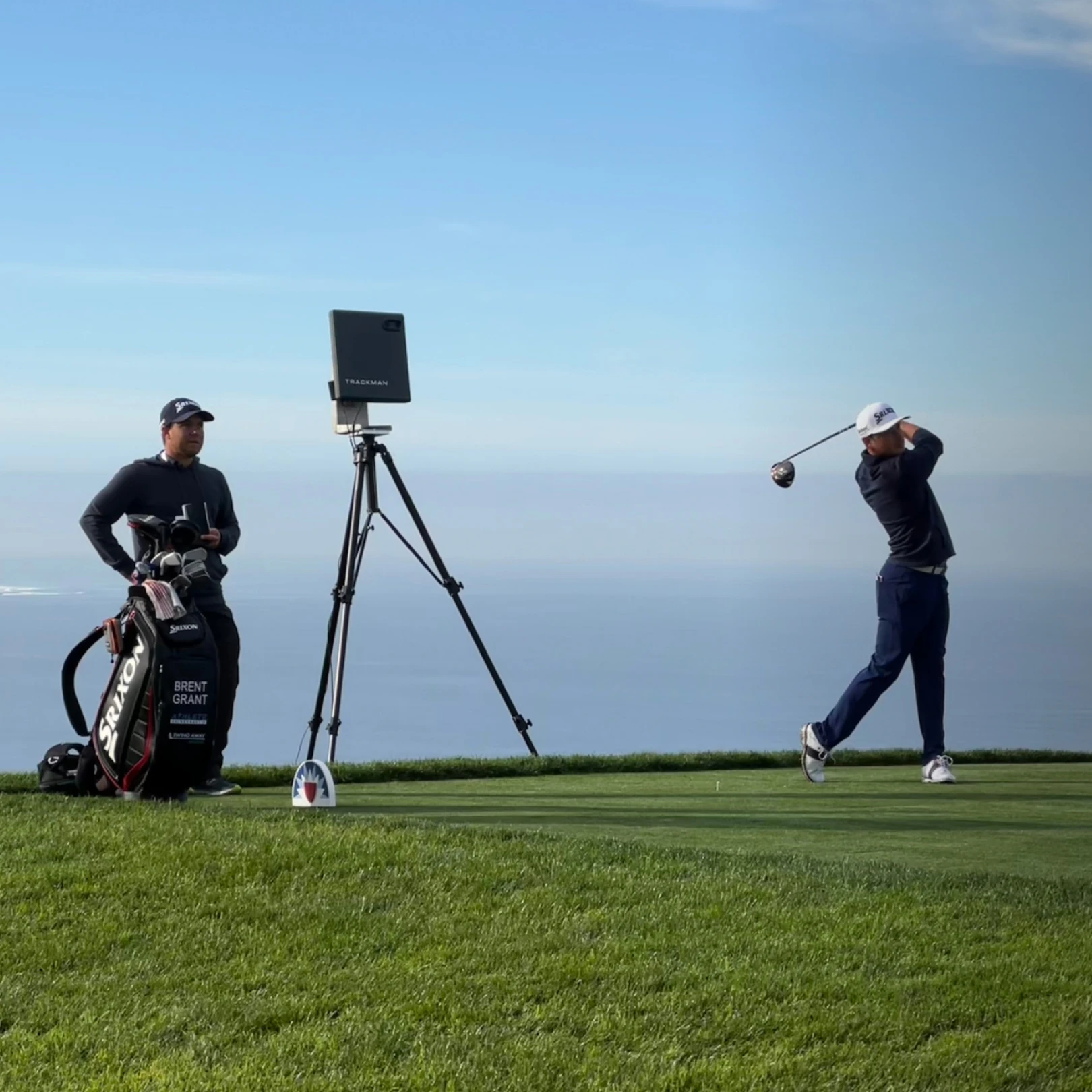 Golfer finishes swing on a sunny course near the water. Caddie stands nearby holding clubs, with a camera set up between them.