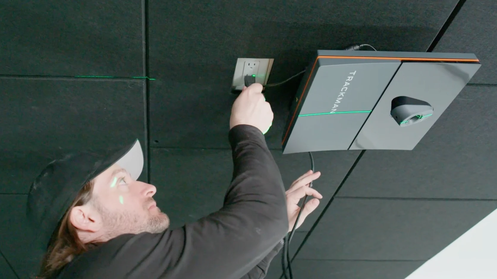 Man wearing a cap installing a Trackman device on the ceiling, plugging it into a power outlet.