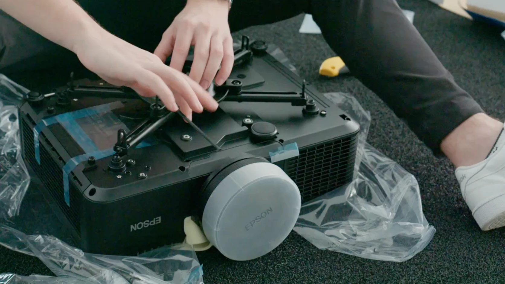 Person assembling an Epson projector, adjusting its stand on a carpeted floor, surrounded by plastic packaging.