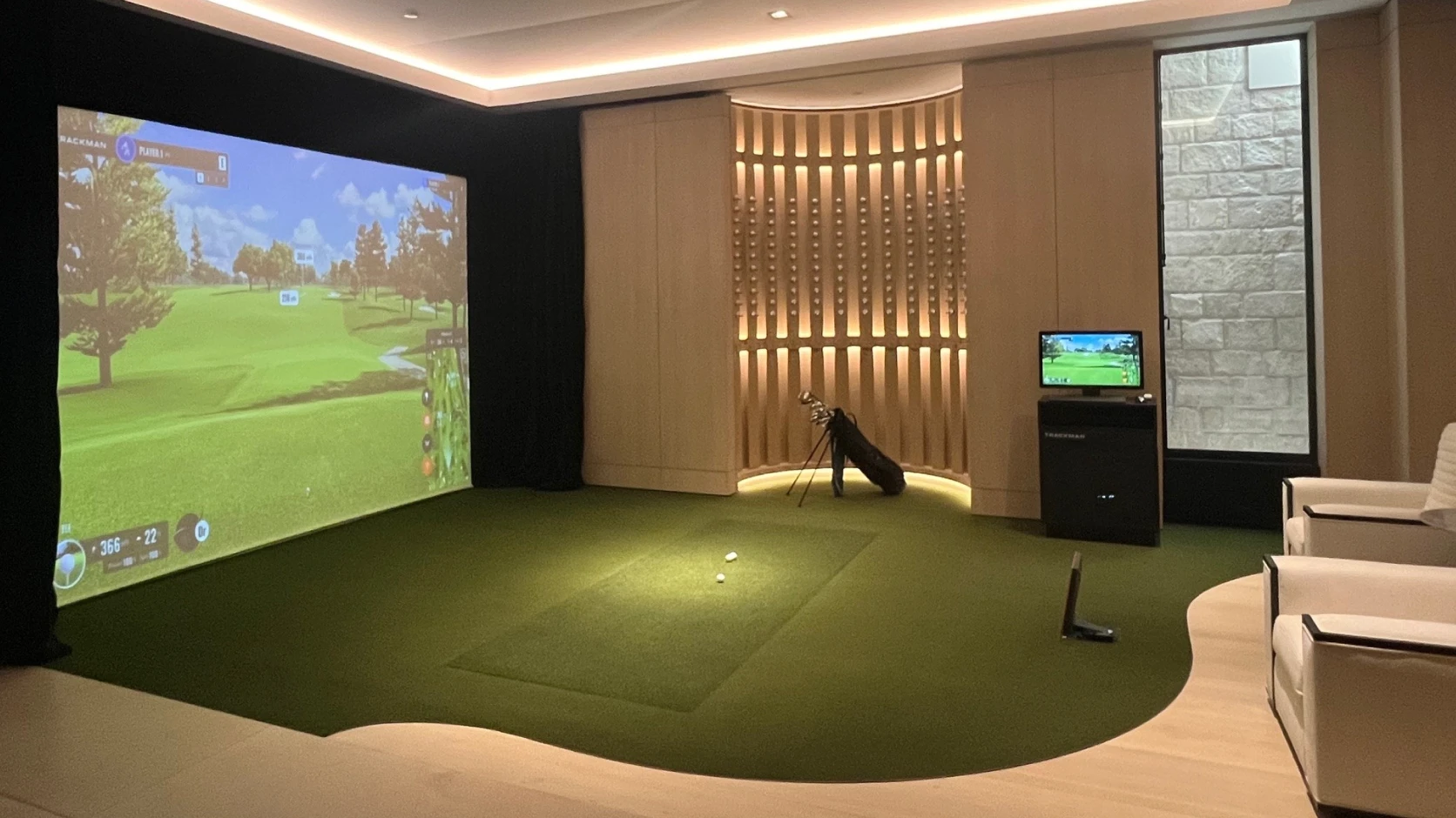 Indoor golf simulator setup with a large screen displaying a golf course, surrounded by golf equipment, chairs, and ambient lighting.