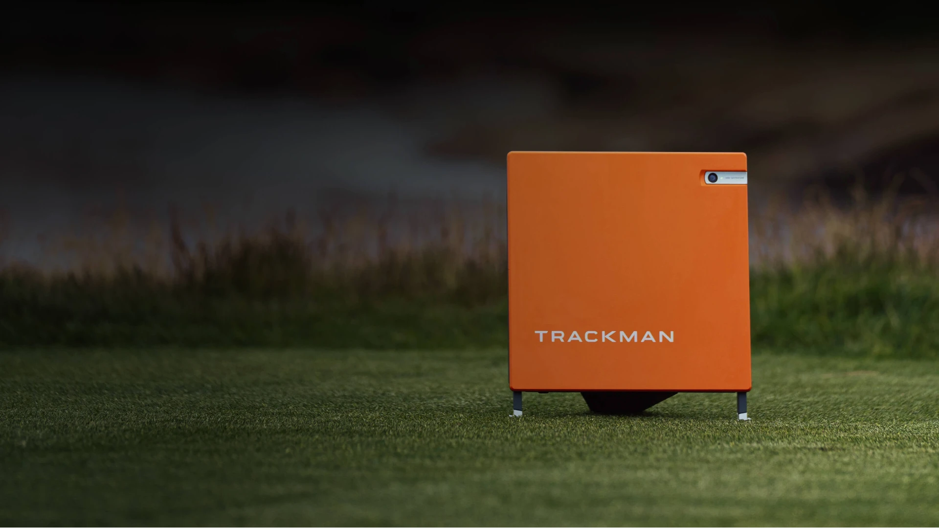Orange TrackMan device on a grassy field, used for sports performance analysis, with a blurred background.