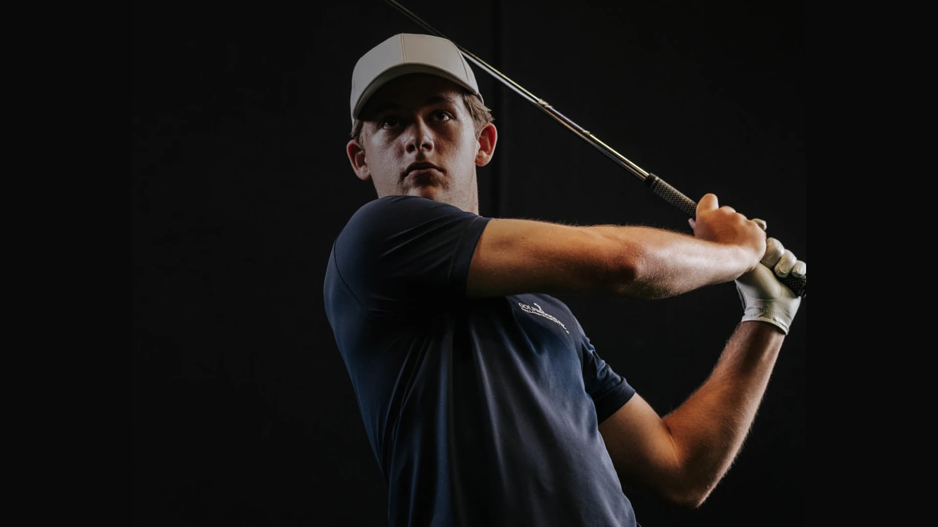 Golfer in a navy shirt and cap swings a club on a dark background, lighting focused on his concentrated expression.