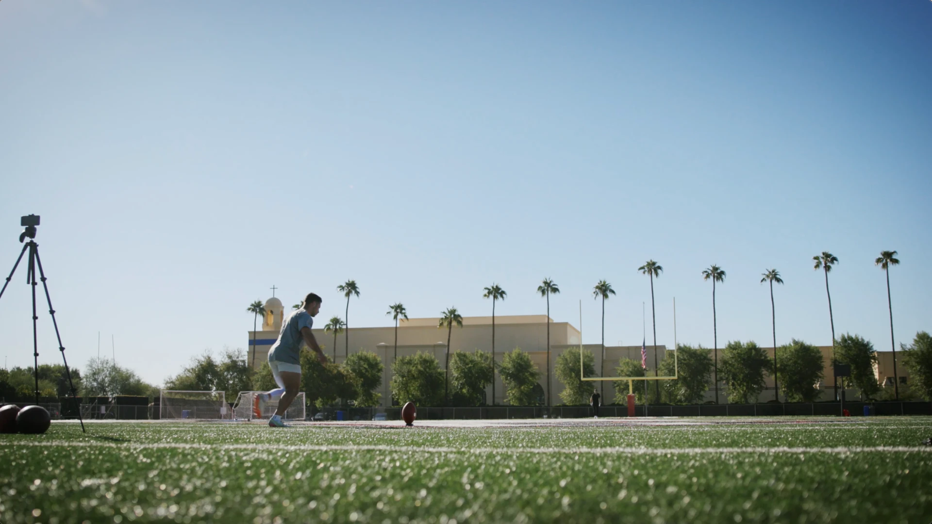 Kicker running to a placed football on a sunlit turf field, tripod camera at left, palm trees and goalposts in the background.