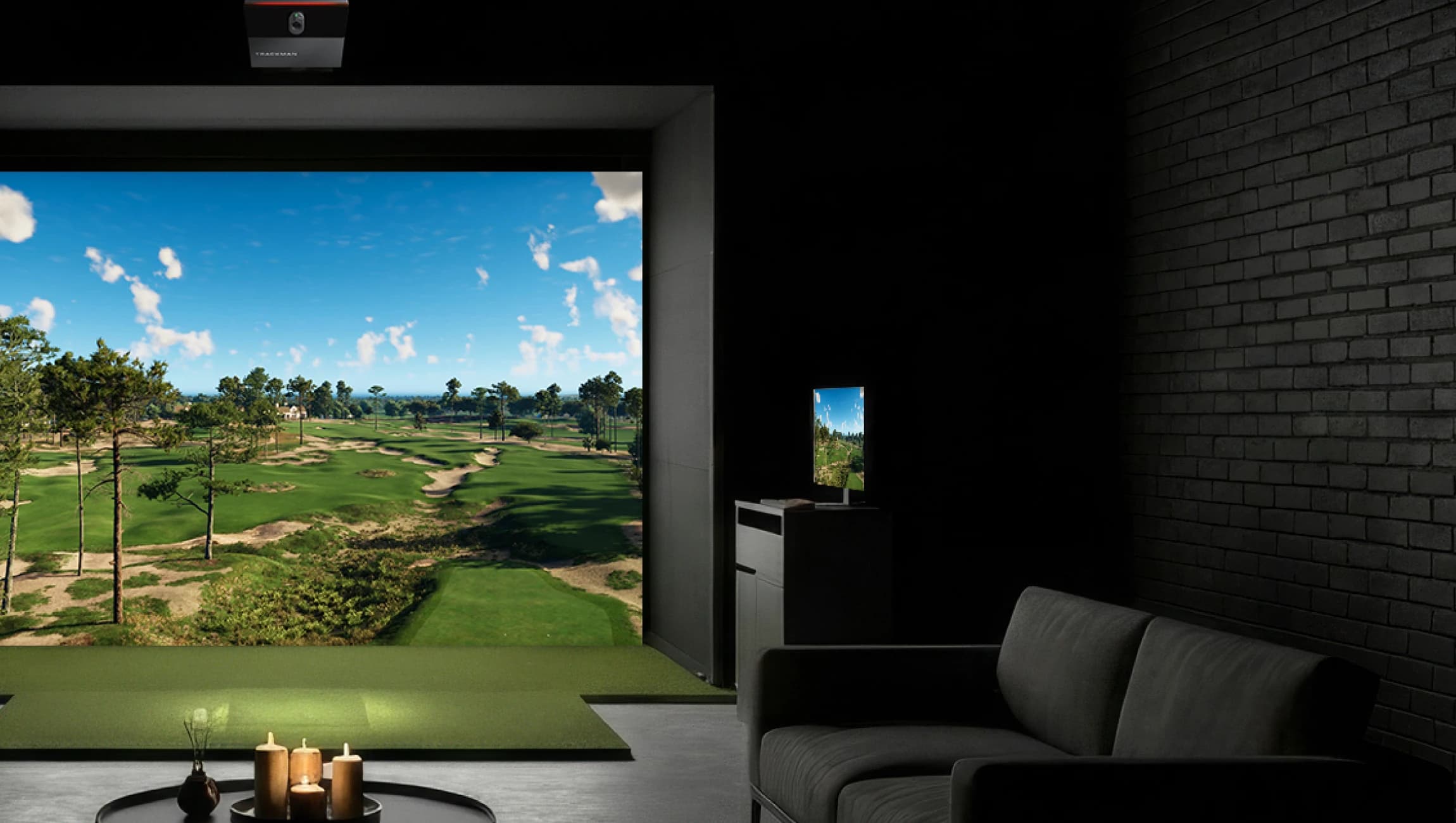 Home golf simulator room with a screen showing a sunny golf course. A sofa and candles in the foreground, dark walls around.