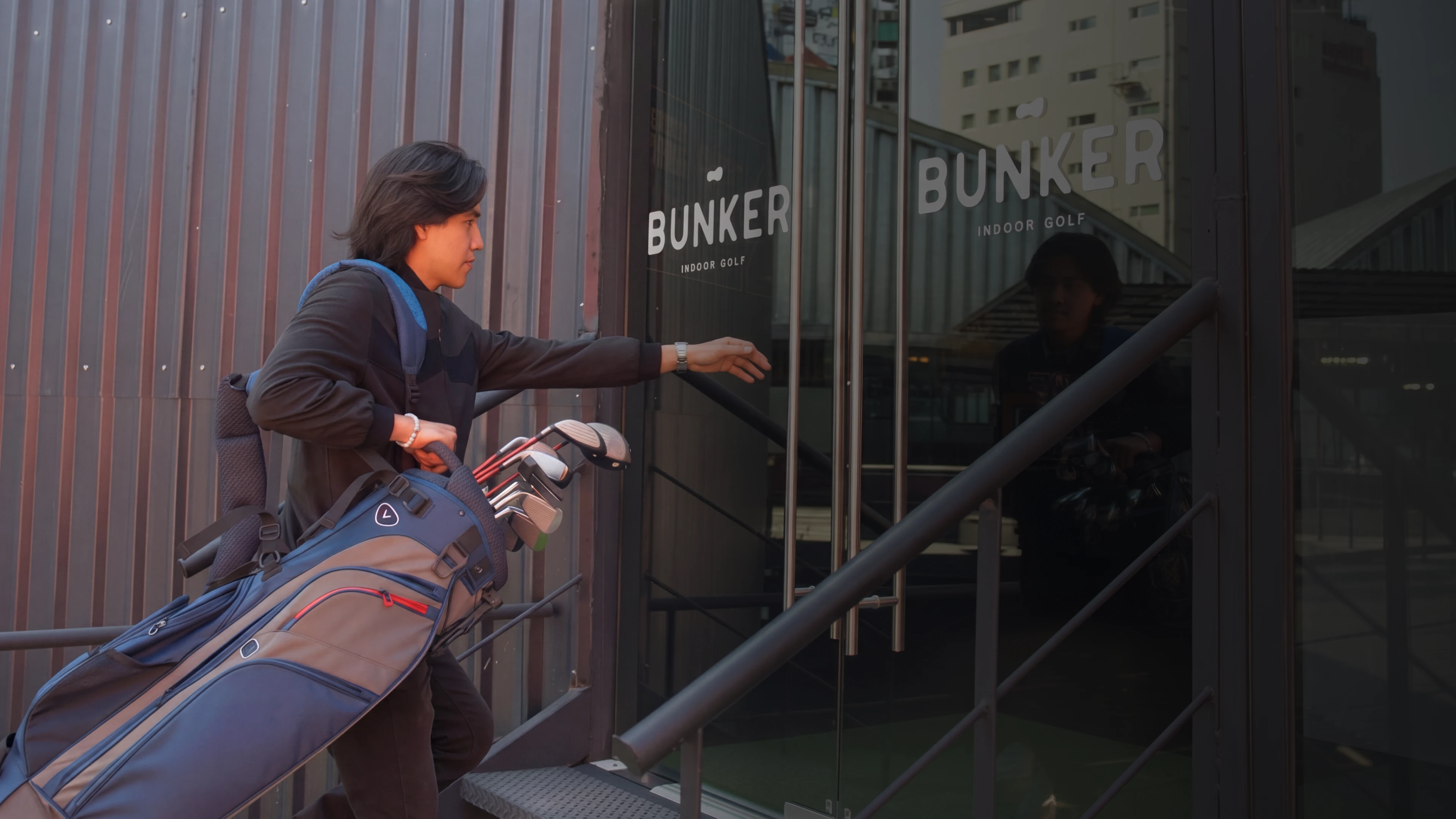 Person carrying golf clubs, reaching for the door of "Bunker Indoor Golf" in an urban setting.