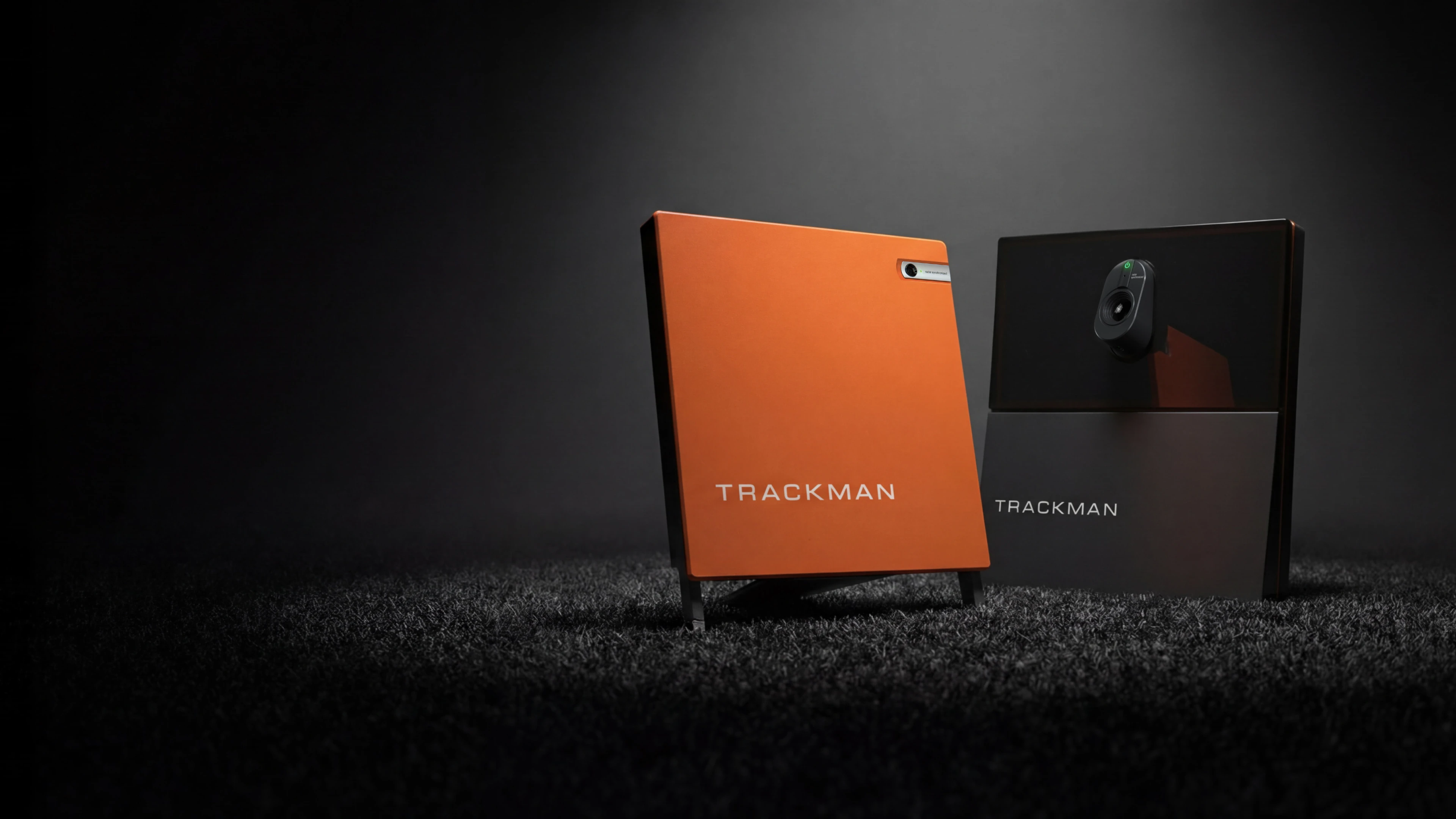 Two TrackMan devices on a dark textured surface, one orange and one black, with sleek, modern designs under spotlight.