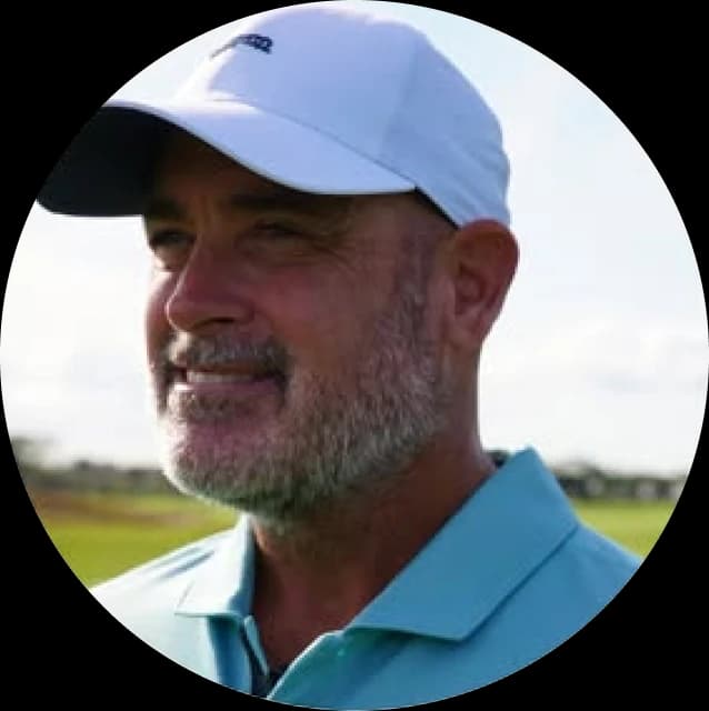 A man with a beard wearing a white cap and light blue shirt is smiling, looking towards the left, with a grassy field in the background.