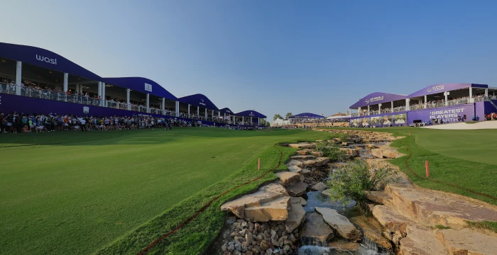 Golf course with a small stream, surrounded by green grass and a large, purple-sheltered spectator area under a clear blue sky.
