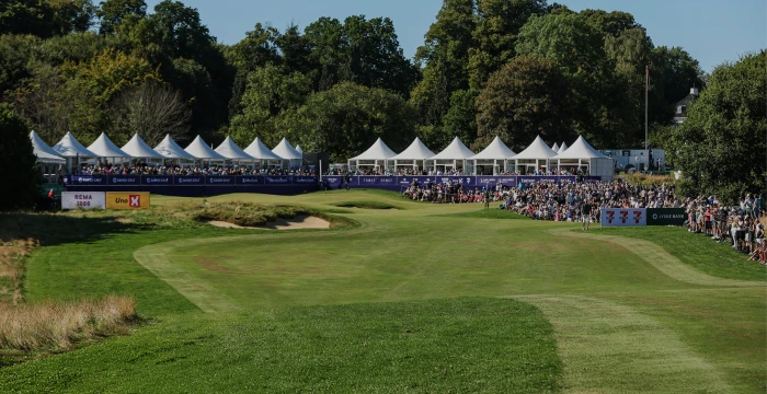 Golf course with a green lawn, surrounded by spectators under white tents, set against a backdrop of trees and a clear blue sky.