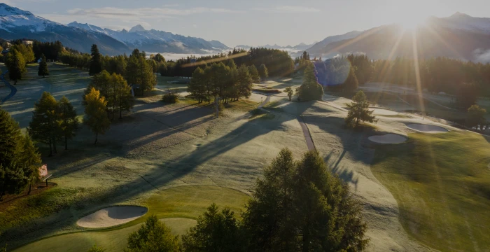 Sunlit golf course surrounded by trees, with mountains in the distance and morning mist, casting long shadows on the grass.