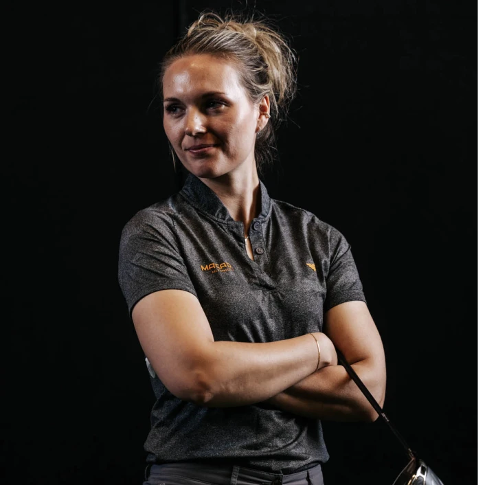 Woman in a gray polo shirt poses with arms crossed, holding a golf club, against a dark background.