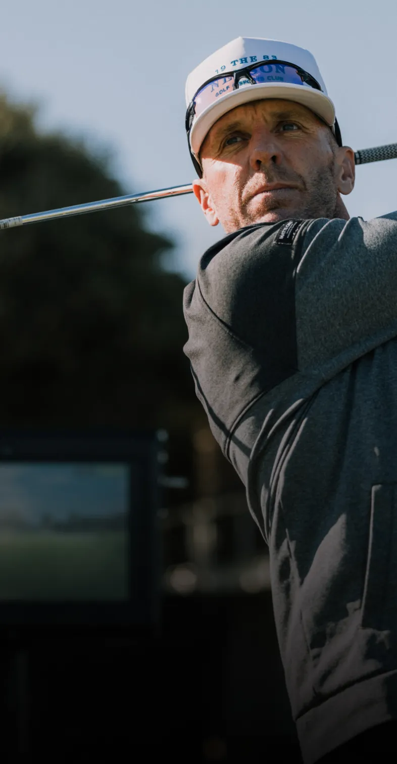 Man in a cap swinging a golf club on a sunny day, with a screen in the background displaying scenery.
