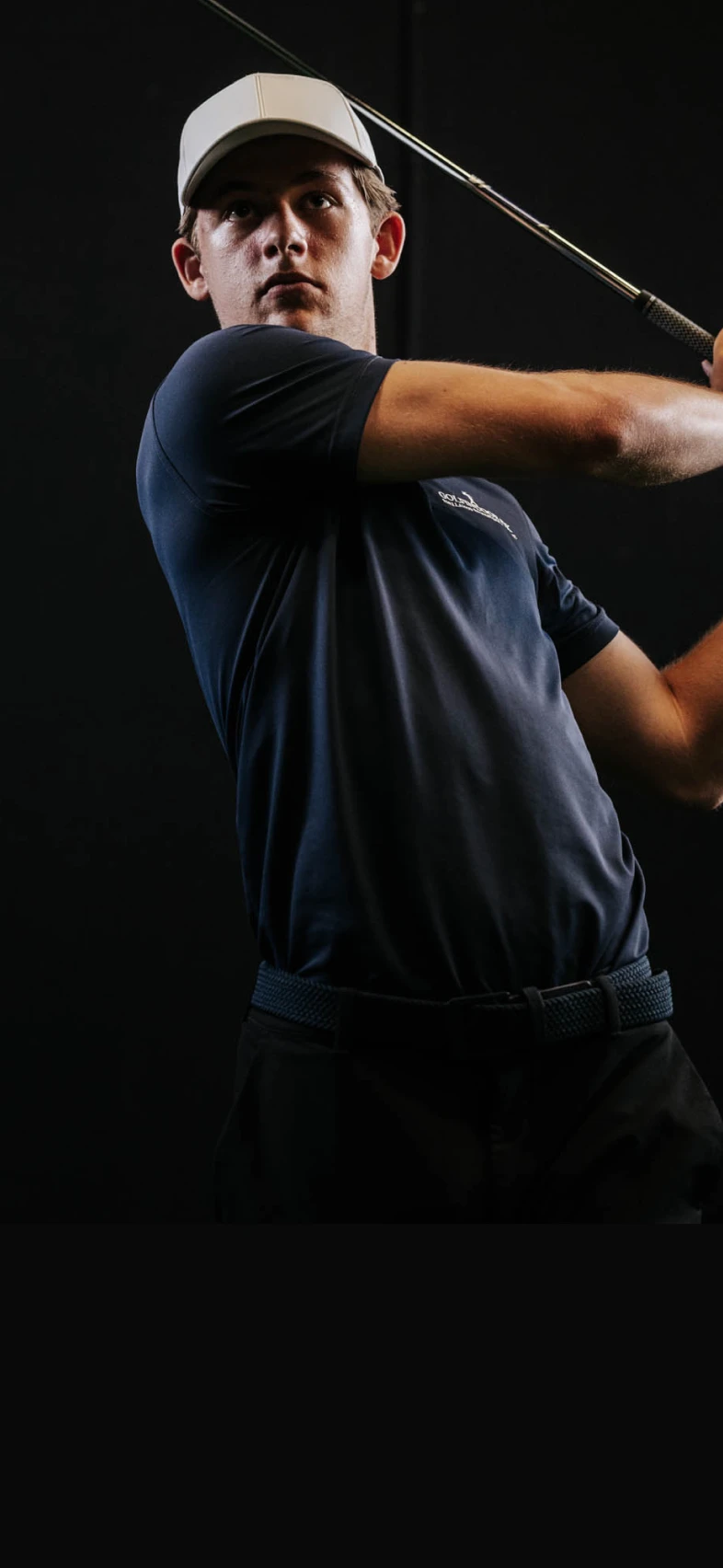 Golfer in a navy shirt and white cap swings a club against a dark background.