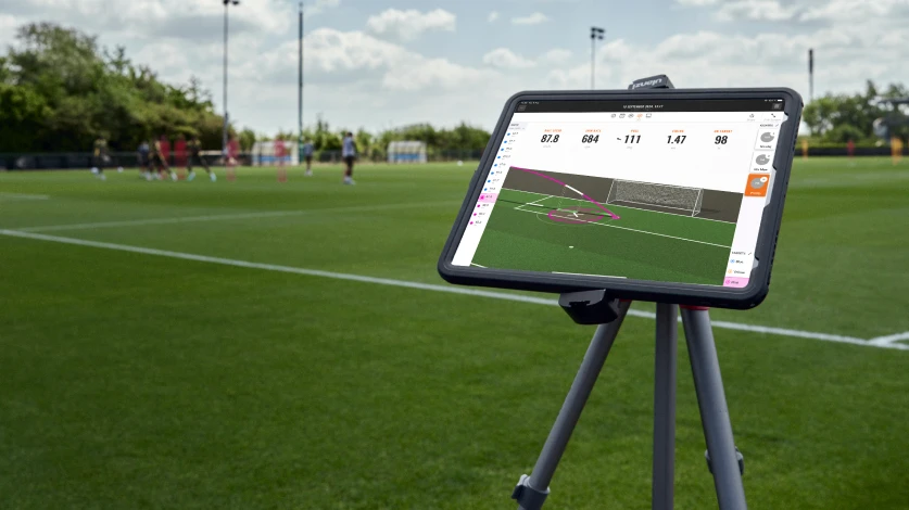 Tablet on tripod showing soccer analytics, focused on a nearby field with players in the background.